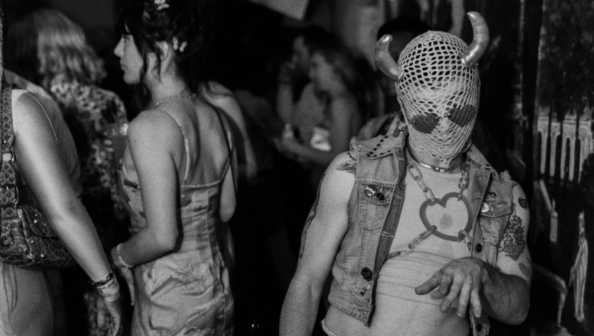 Person in horned mask and leather outfit poses at a crowded indoor party.