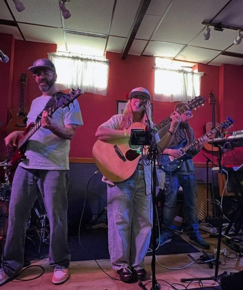 A five-piece band performs on stage with guitars, drums, and keyboards in a red-walled room.