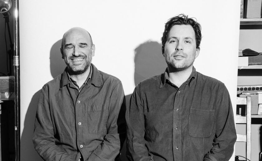 Two men in button-up shirts sit side by side, one smiling with eyes closed, in a studio setting.
