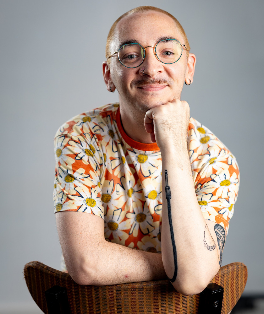 Person with glasses and floral shirt smiling, sitting backward on a chair, arm resting on chair.