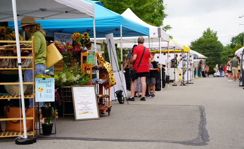 Outdoor market with people browsing vendor booths and flower bouquets displayed on tables.