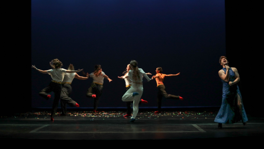 Six performers dance and sing on stage under dramatic lighting, with confetti on the floor.