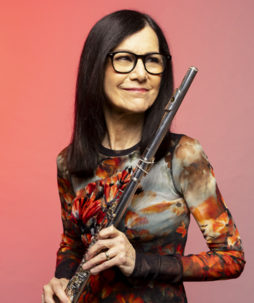 Woman with long dark hair and glasses holding a flute, wearing a floral patterned top.