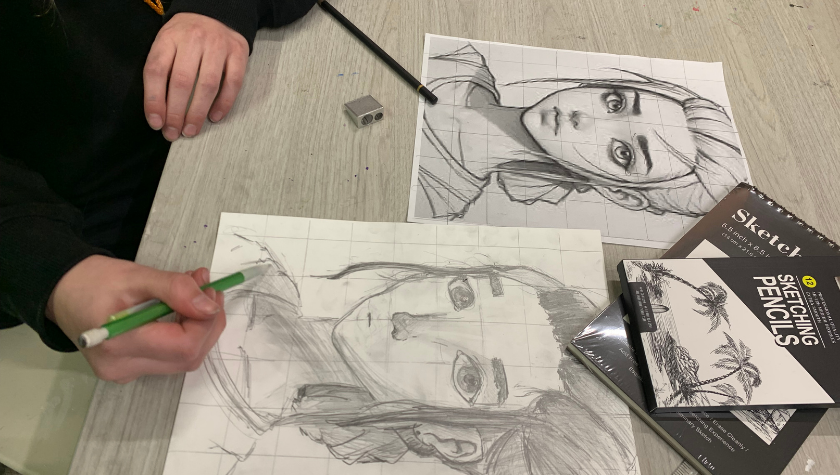 Person drawing a pencil portrait on grid paper with reference image and sketching supplies nearby.