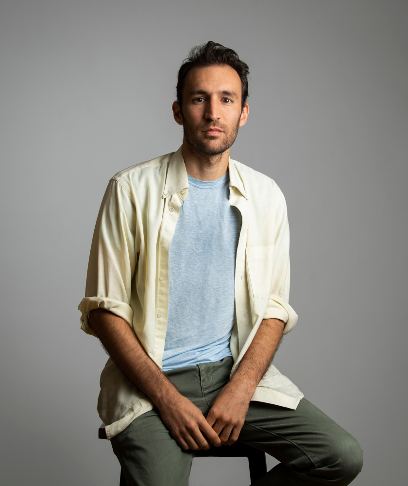Man in casual clothes sitting on a stool against a plain grey background.