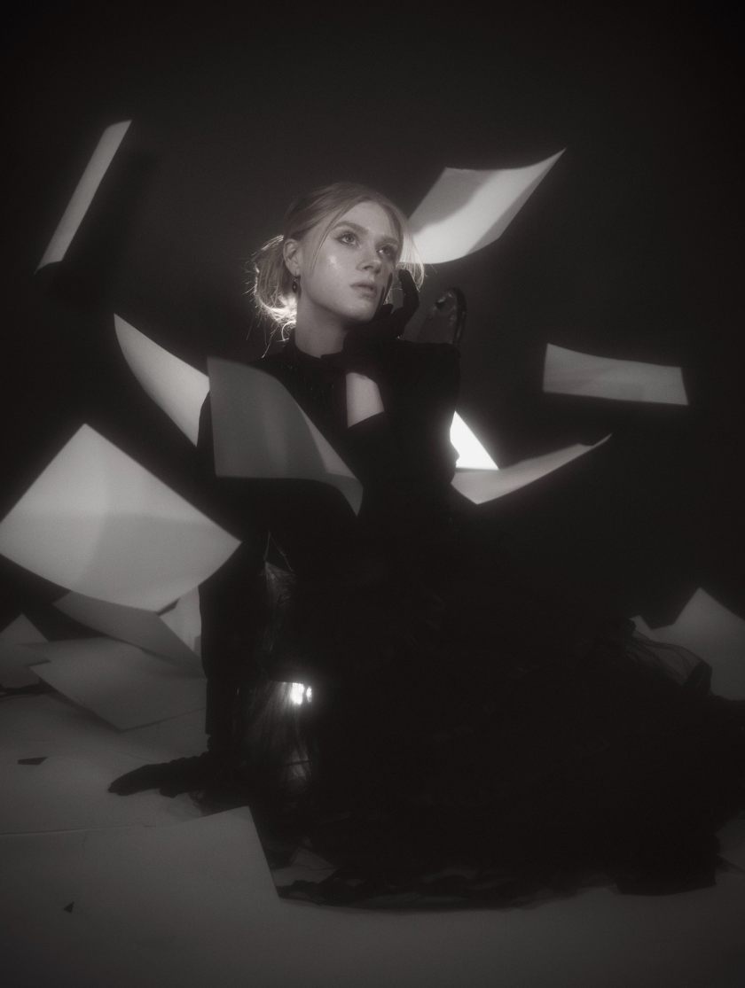 A woman in a dark dress sits on the floor as papers dramatically float around her.