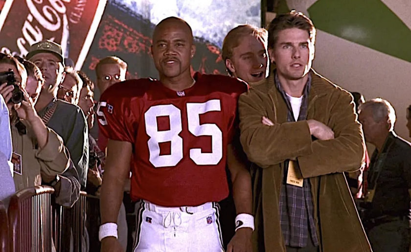 A football player in a red jersey stands beside a man in a brown jacket amid a crowd.