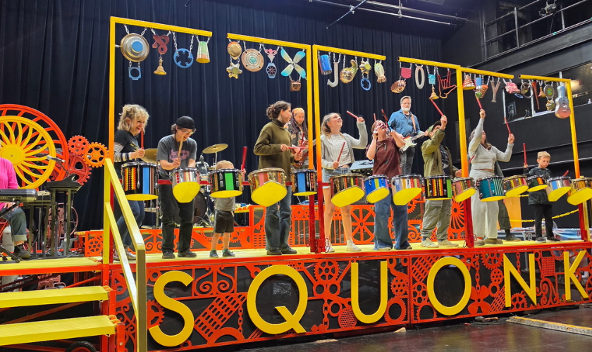 A band performs on a colorful stage with drums and hanging objects, SQUONK displayed below.