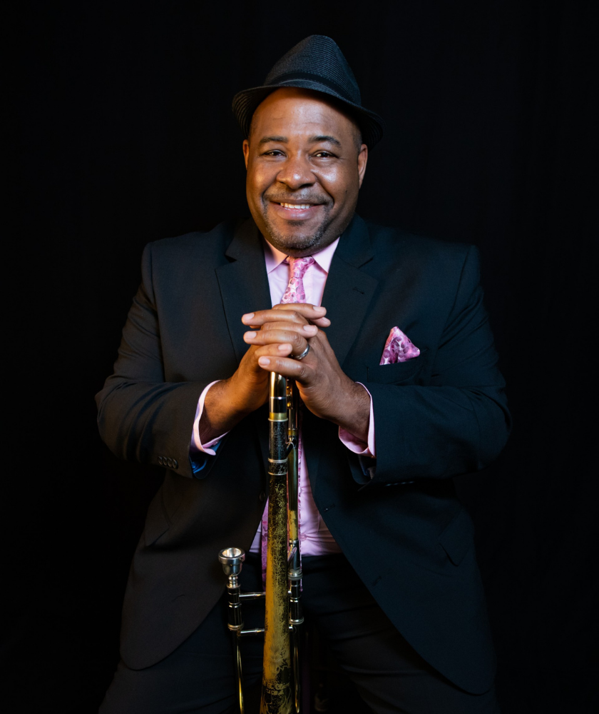Man in a suit and hat smiling, holding a trombone, seated against a black background.
