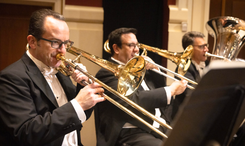Three trombone players in tuxedos performing in an orchestra.