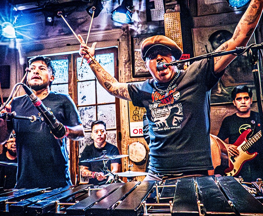 A lively band performs in a colorful room, with one musician playing a xylophone and singing passionately, arms raised.