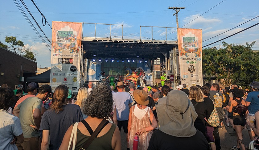 A crowd gathers outdoors in front of a stage where a band is performing at a street festival.