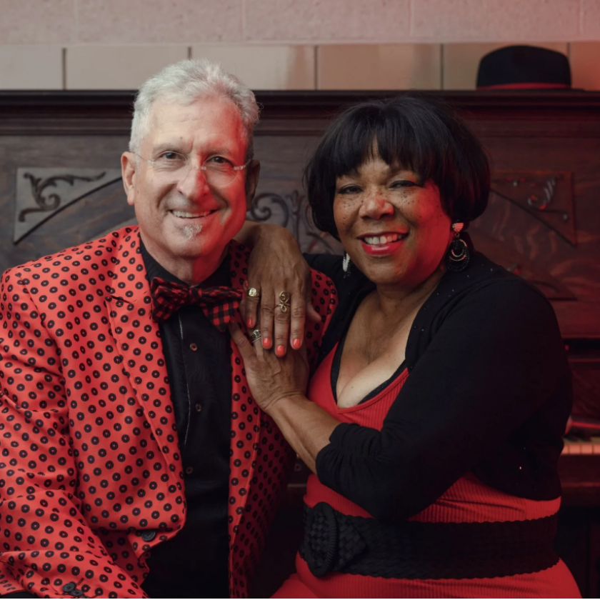 Smiling man in a red patterned suit and woman in a red dress sit together by a piano.