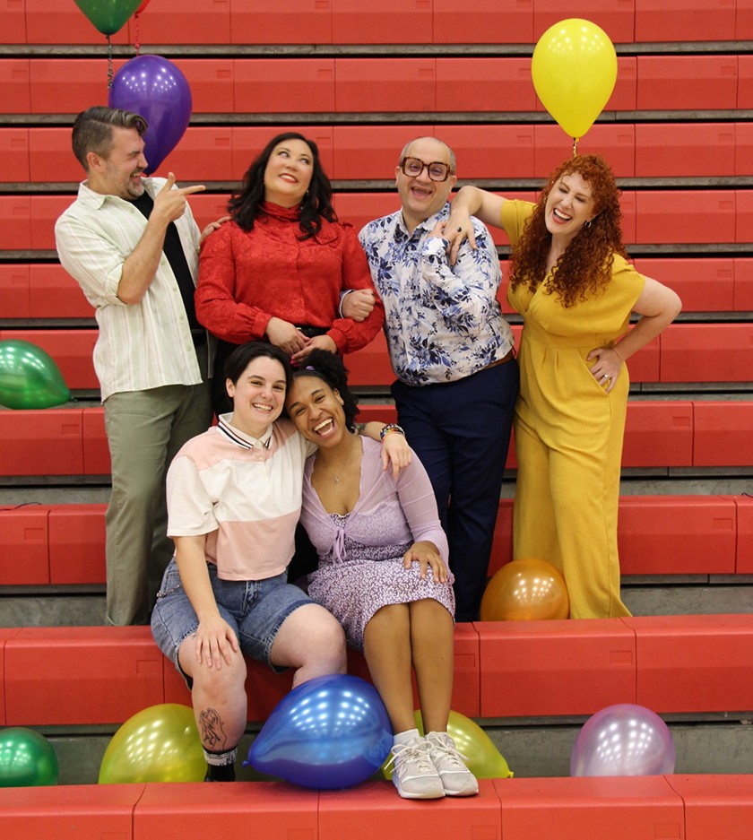 Six smiling adults pose together on red bleachers with colorful balloons. Two people sit in front, while four stand behind them. They appear happy and playful in casual and bright clothing.
