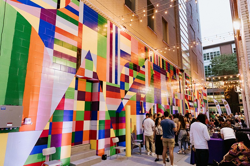 A lively alleyway event with people gathered near a bar, set against a colorful geometric mural on a tall brick wall, under string lights in the evening.