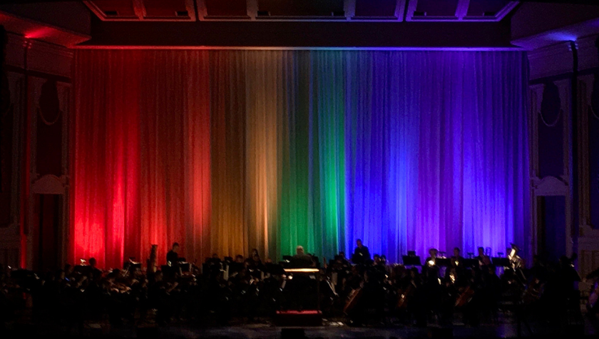 A symphony orchestra plays onstage as rainbow lights illuminate the curtain backdrop.