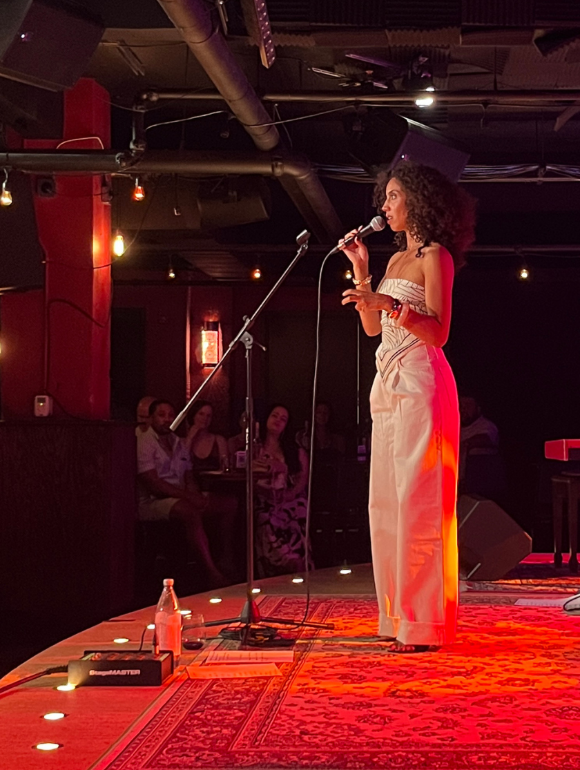 Woman sings on stage with musicians; red lighting and audience visible in a cozy venue.