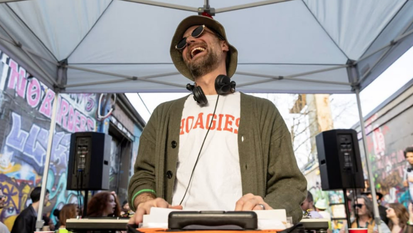 DJ wearing sunglasses and a bucket hat smiles while performing at an outdoor event.