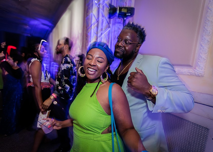 Two people smile and pose for the camera at a lively indoor party, with purple and blue lighting. Other guests are dancing and mingling in the background. The woman wears a green dress and blue hat, and the man wears a light-colored suit.