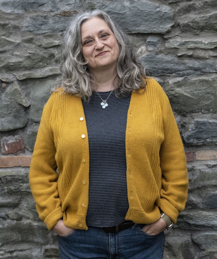 A person with long, wavy gray hair stands in front of a stone wall, smiling slightly. This person has their hands in their jean pockets and is wearing a yellow cardigan.