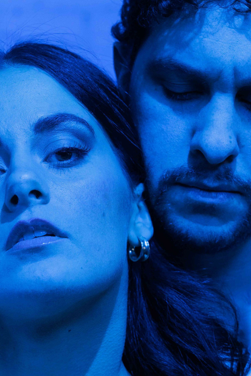A moody close up of two people in blue lighting with their heads together. One is looking at the camera, the other is looking down.