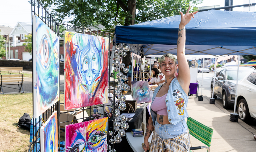 Smiling person raises arm at outdoor art booth with colorful abstract paintings and decorations.