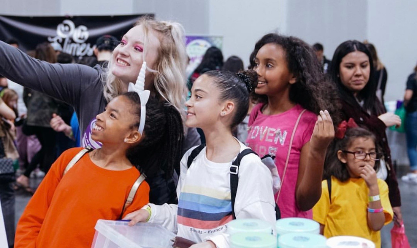 Group of kids and an adult smiling and posing together at an indoor event or convention.