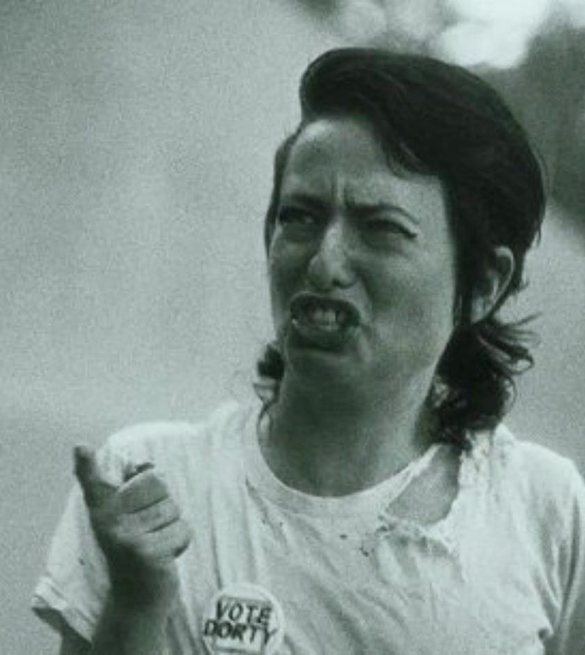 Person in a torn shirt making a grimace, wearing a VOTE DORT button.