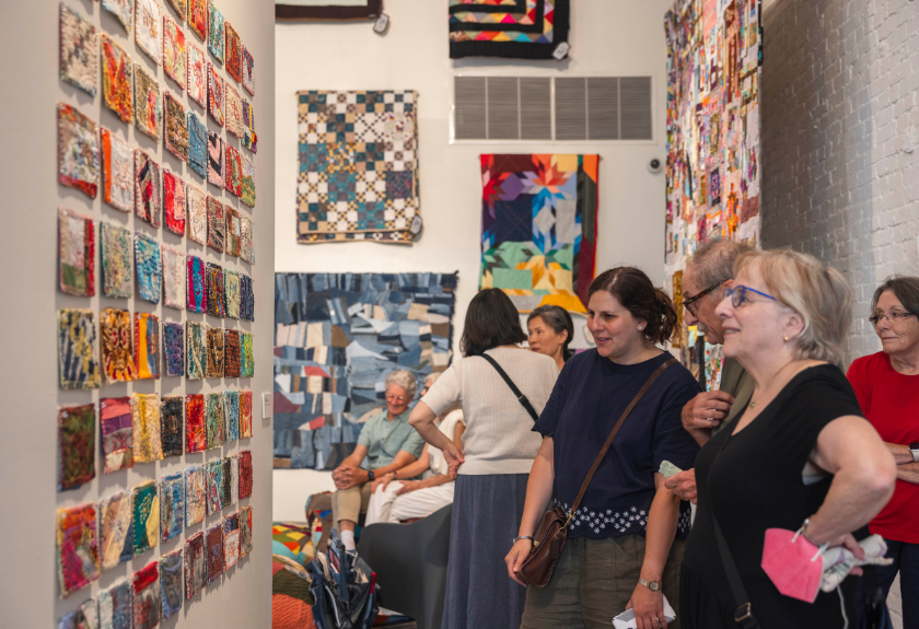 People observe colorful quilts and fabric squares displayed on gallery walls.