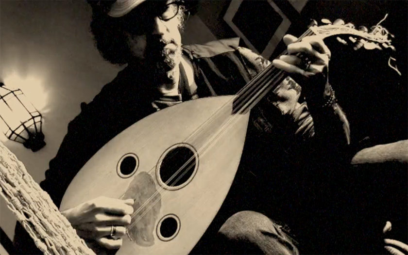 A person wearing glasses and a cap plays an oud, a stringed musical instrument, in a dimly lit room. The image is in sepia tones, creating a vintage and artistic atmosphere.