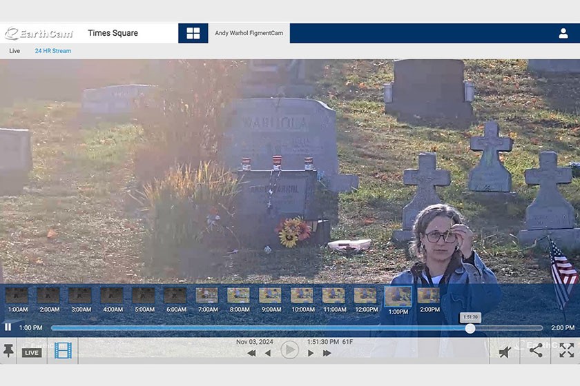 A woman stands in front of a gravestone and several crosses in a cemetery during daytime. She wears glasses and holds a phone, while sunlight shines from the upper left of the image. A video timeline is visible at the bottom.