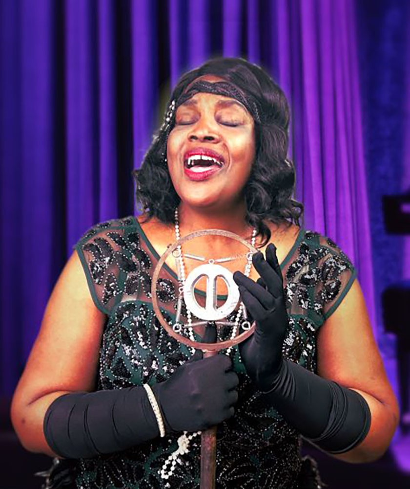 A woman in a vintage, beaded black dress and gloves sings passionately into an old-fashioned microphone, standing in front of purple curtains.
