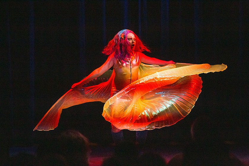 A performer with colorful hair dances on stage, holding large, translucent, fan-like wings that glow with orange and yellow light against a dark background.