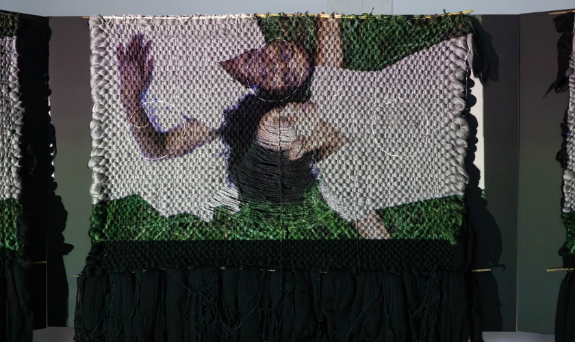 A woven textile shows two people lying on grass with one arm raised, seen from above.