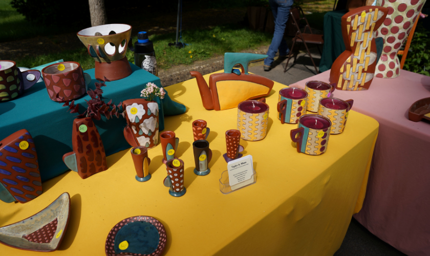 Various colorful pottery pieces are displayed on a yellow tablecloth at an outdoor market.