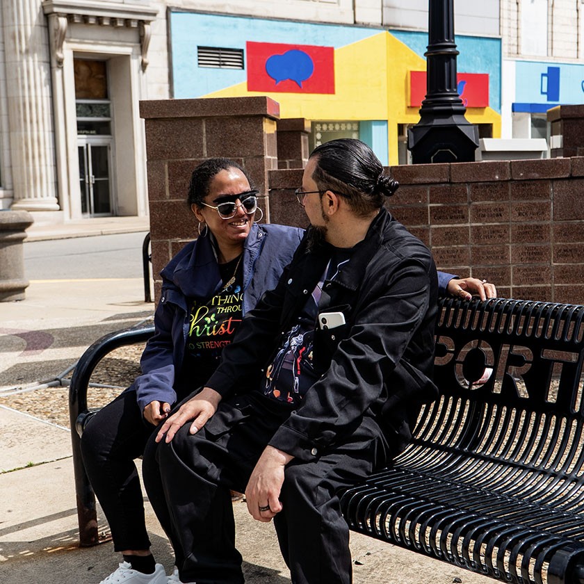 Two people sit on a black metal bench outdoors, engaged in conversation and smiling. Colorful geometric art is visible on the building in the background.