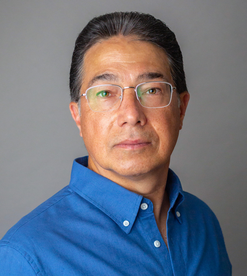 Man with glasses and gray hair wearing a blue shirt, looking at the camera, gray background.