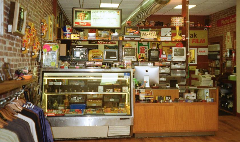 Vintage store counter with snacks, electronics, and Kodak film signs, surrounded by brick walls.