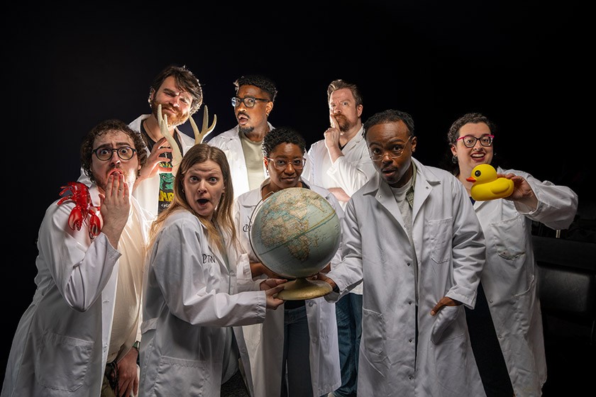 A group of seven people in lab coats pose dramatically with quirky props, including a globe, a rubber duck, a lobster, a can, and antlers, against a dark background. They have playful and surprised expressions.