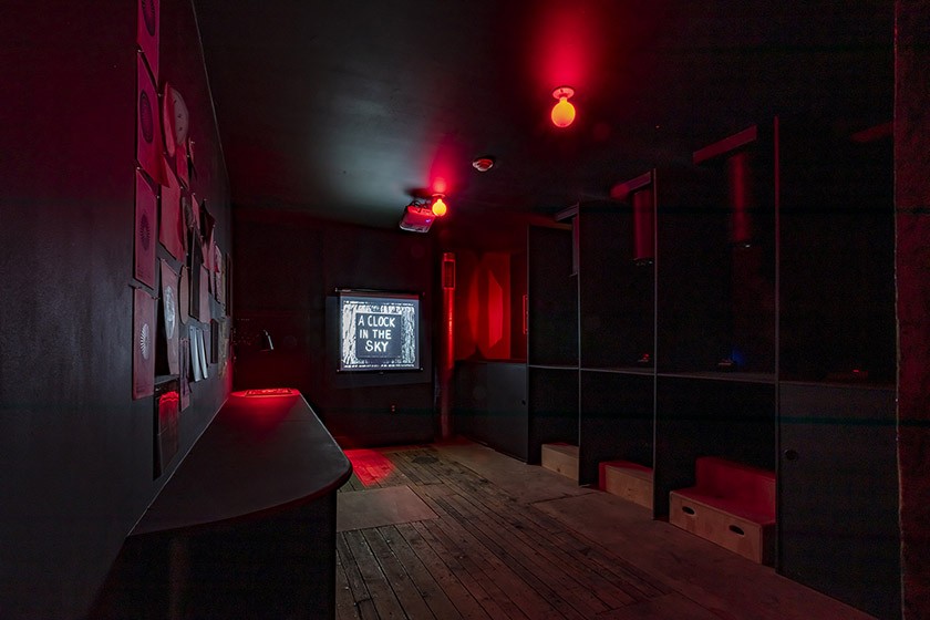 A dimly lit room with dark walls illuminated by red lights. A bulletin board with papers is on the left, and a screen at the back displays the text A LOCK IN THE SKY. Wooden floors and benches are visible