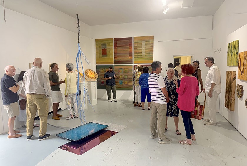 A group of people mingle and view colorful modern artworks in a bright, white-walled gallery. Sculptures and paintings are displayed on the walls and floor, creating a lively and engaging atmosphere.