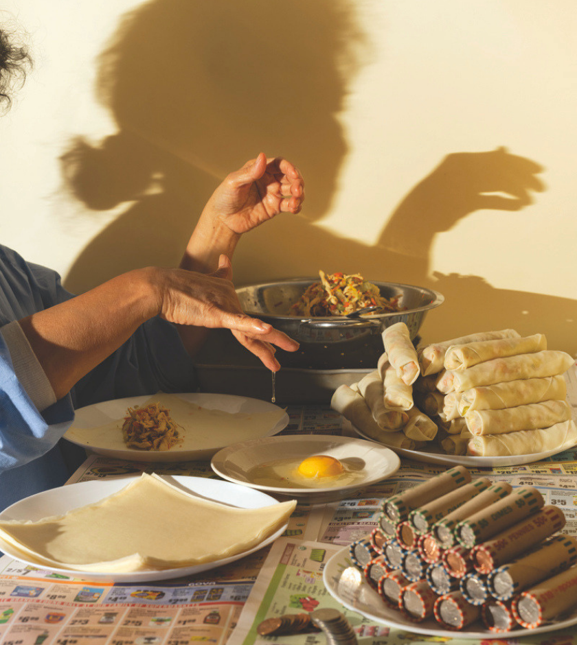 Person making spring rolls, with wrappers, egg, filling, and rolls on a table in warm light.