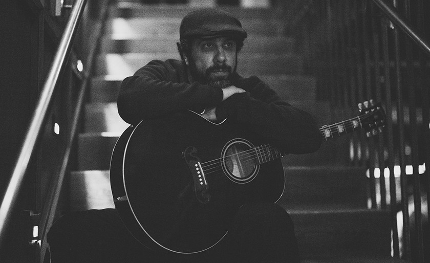 A man in a flat cap sits on stairs, holding an acoustic guitar and resting his arms on it, gazing thoughtfully ahead. The photo is in black and white.