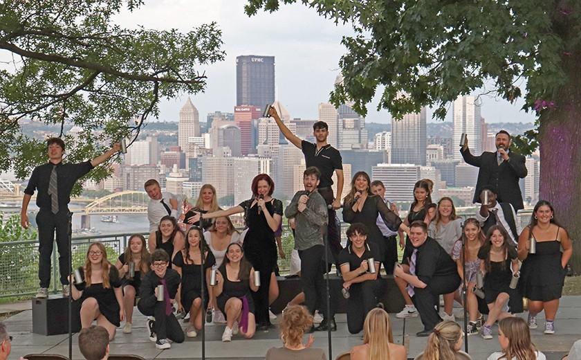 A group of young performers in black attire pose energetically on an outdoor stage, holding microphones, with a city skyline and bridges visible in the background under a cloudy sky.