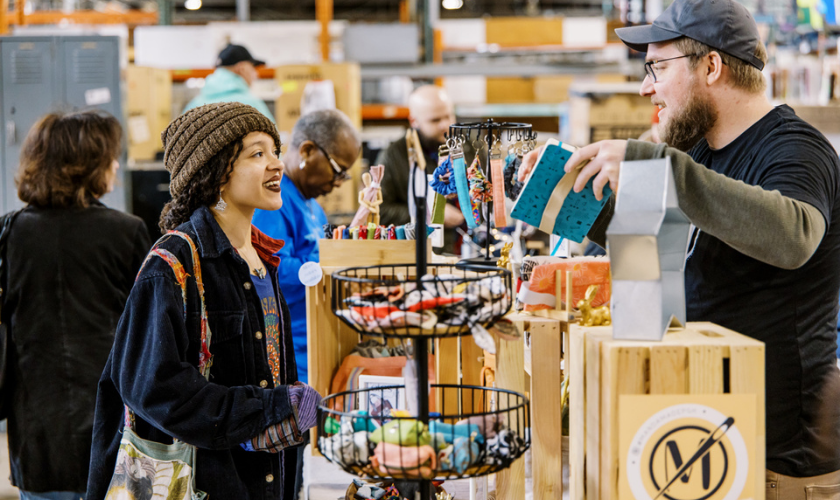 Woman smiles at vendor at a craft market booth with handmade items on display.