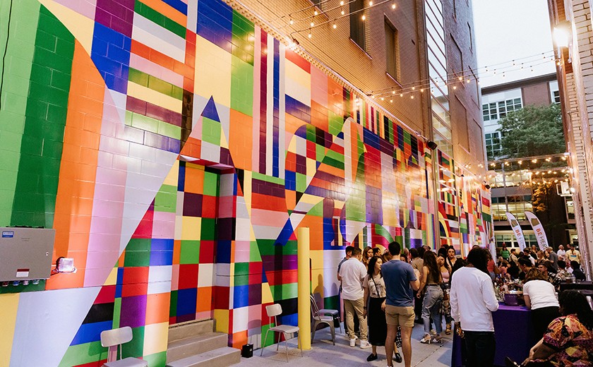 People gather in an outdoor alley decorated with colorful, geometric mural art on the walls and string lights overhead, creating a lively, festive atmosphere.