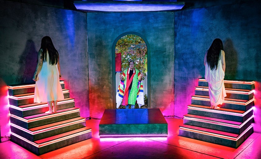 A colorful stage scene features two people in white dresses standing on opposite staircases with glowing neon lights, and a central figure in a multicolored robe standing in an arched doorway at the back, arms raised.