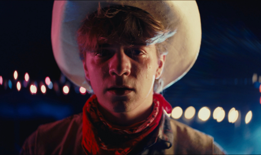 Young person in a cowboy hat and red bandana stands in front of blurred night lights.