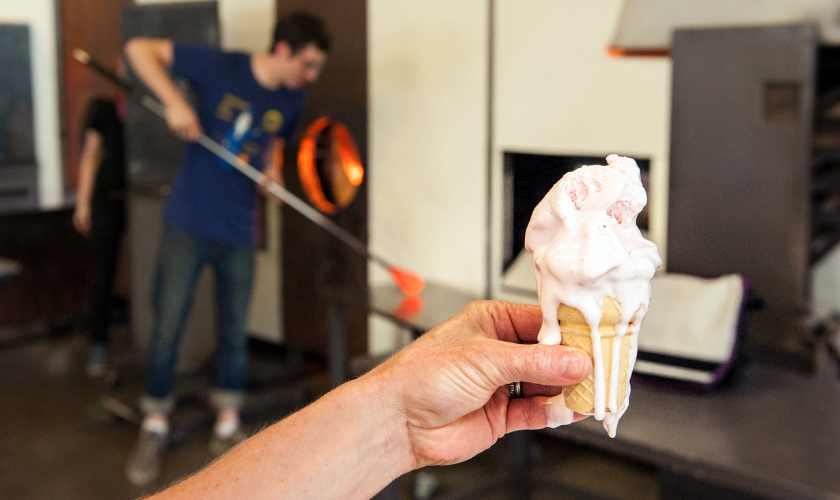 A hand holds a melting ice cream cone; a person in the background works with molten glass.