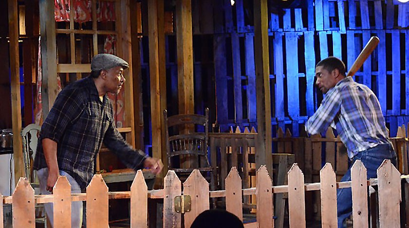Two men face each other intensely across a wooden fence on a stage; one man holds a baseball bat threateningly, while the other stands defensively. The set resembles a rustic yard with wooden pallets and blue lighting.